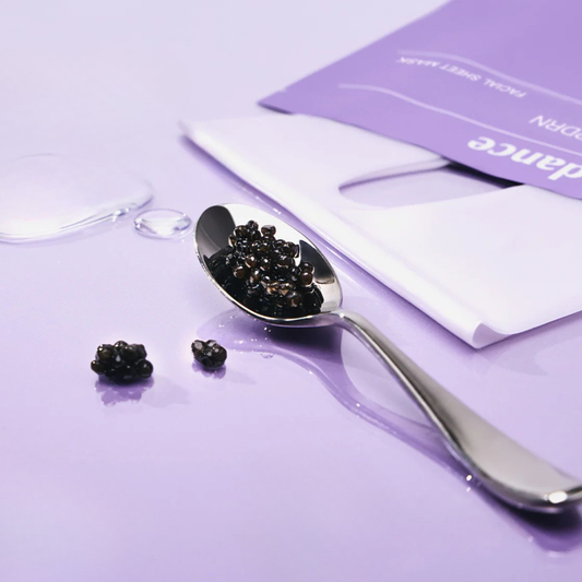 Black pearl caviar on a silver spoon with a few pearls scattered on a glossy lavender surface, next to a BIODANCE Rejuvenating Caviar PDRN Real Deep Mask.