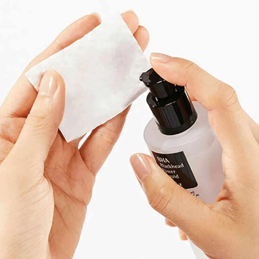 Hands holding a white cotton pad and COSRX BHA Blackhead Power Liquid with a black cap on a white background