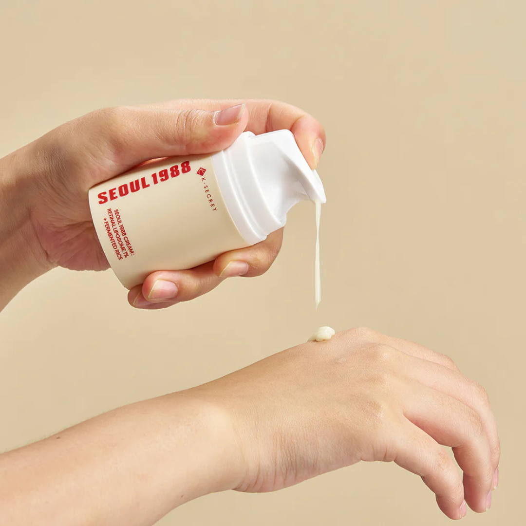 Hand holding a Skincare bottle K-SECRET Seoul 1988 Cream: Retinal Liposome 1% + Fermented Rice skincare product with cream being applied to skin on a beige background. Pressing cream out of bottle onto back of a hand