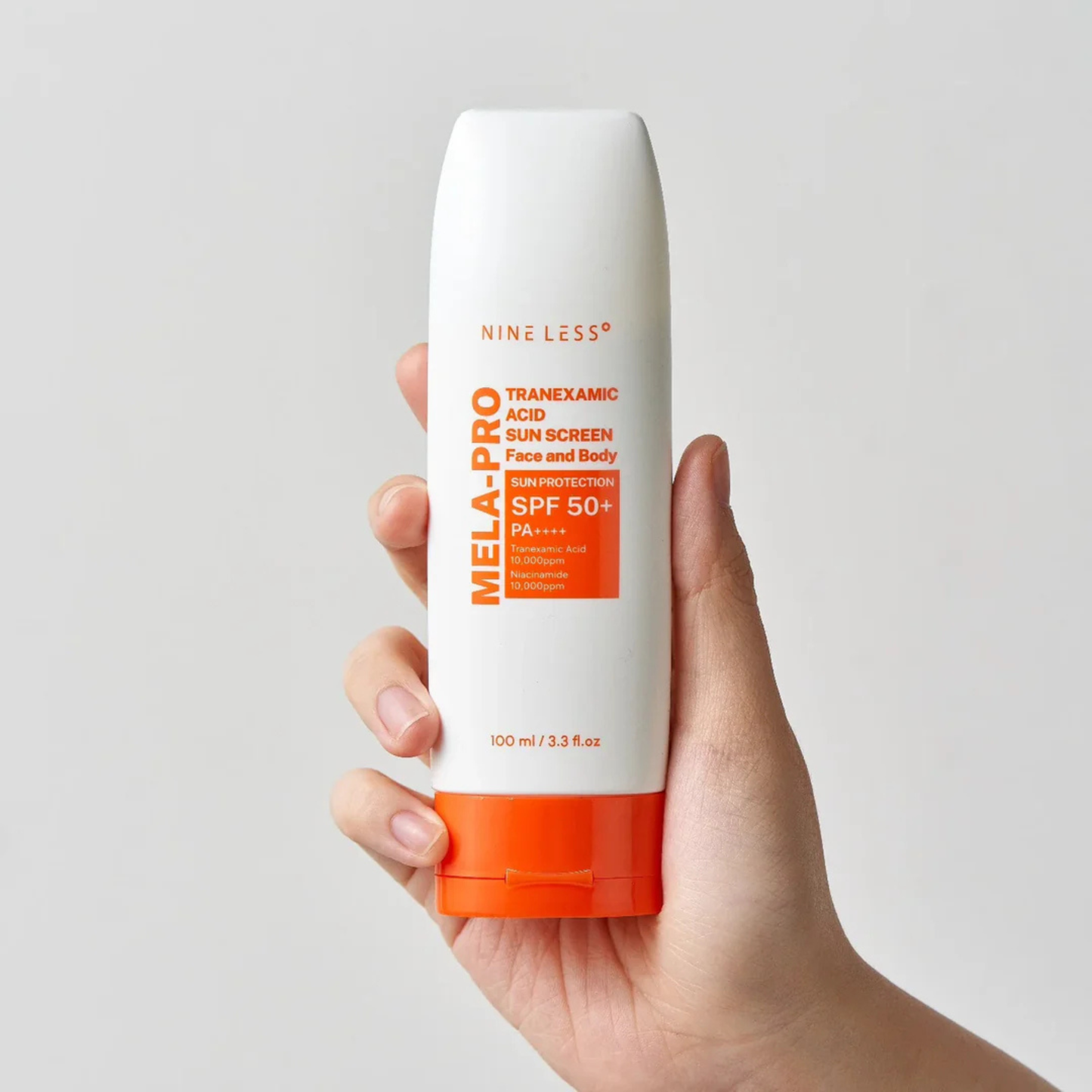 Hand holding a bottle of NINE LESS Mela-Pro Tranexamic Acid Sun Screen with a white and orange design on a grey background