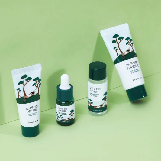 Four skincare products from the ROUND LAB Pine Calming Cica Trial Kit are arranged on a light green background. The set includes a white and dark green hand cream tube, a clear serum bottle with a white dropper, a clear toner bottle with a dark green cap, and a white and dark green body cream tube.