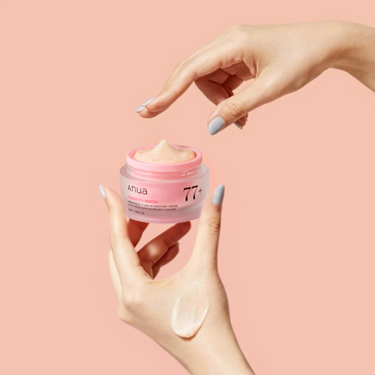 Hand holding a pink jar of Anua Peach 77+ Niacin Enriched Cream with 'Anua' branding on a light pink background