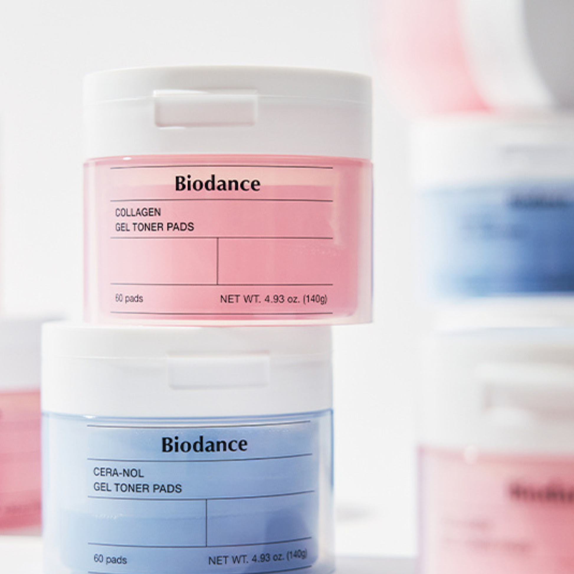 Two Biodance brand jars of collagen and cera-nol gel toner pads stacked vertically on a white surface, with a soft, blurred background showing additional pink and blue jars.