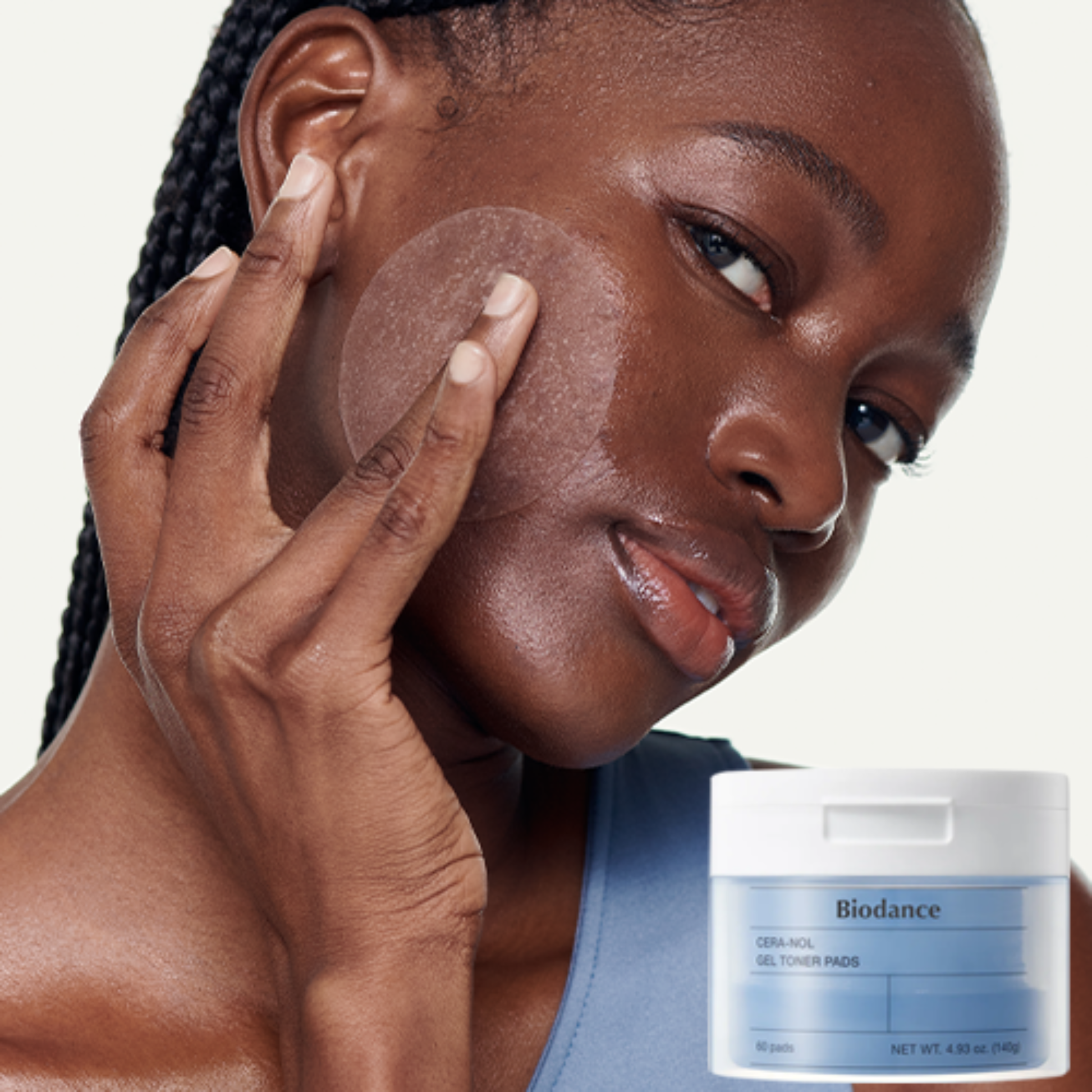 A woman with braided hair applies a translucent round gel toner pad to her cheek, with a white and blue Biodance Gel Toner Pads jar shown in the lower right corner against a light off-white background.