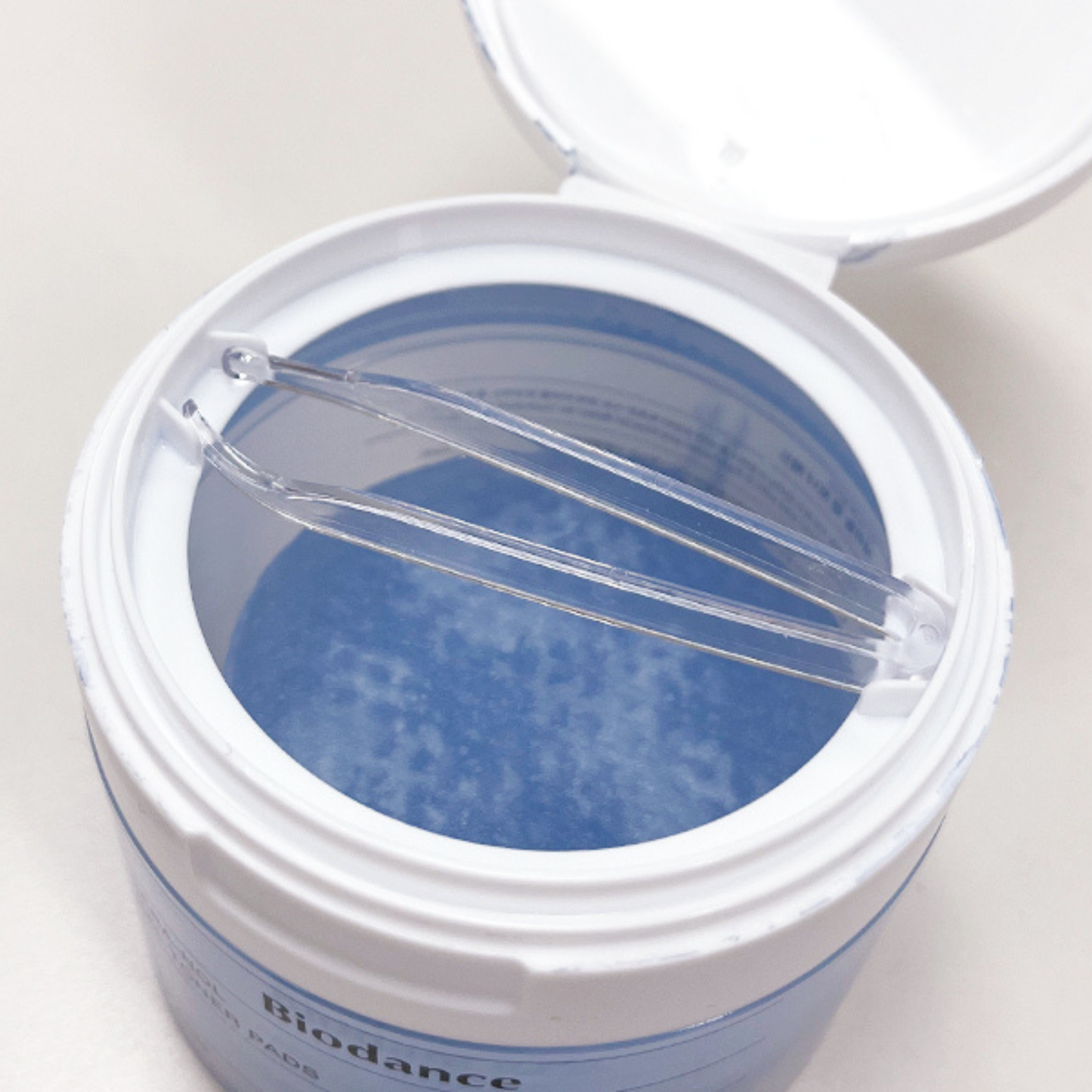 An open white cylindrical container showing Biodance Cera-Nol Gel Toner Pads, holding a pair of clear plastic tweezers. The label at the bottom reads 'Biodance'.