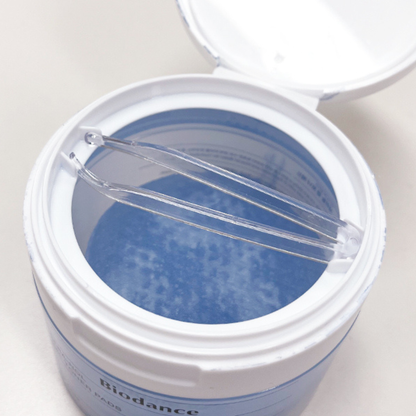 An open white cylindrical container showing Biodance Cera-Nol Gel Toner Pads, holding a pair of clear plastic tweezers. The label at the bottom reads 'Biodance'.