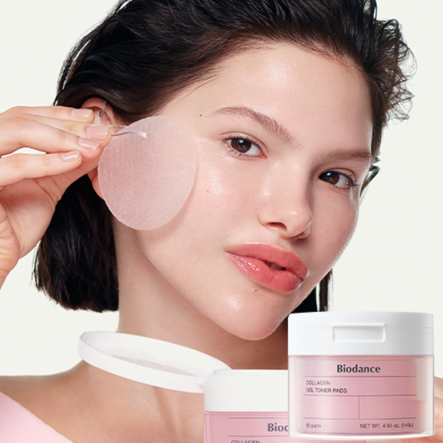 A woman applies a translucent pink gel Biodance Collagen Gel Toner Pad to her cheek. Two jars of Biodance Collagen Gel Toner Pads are shown in the foreground against a clean white background.