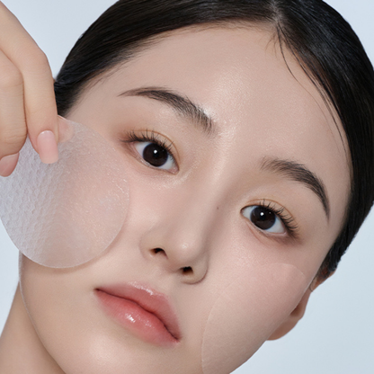 Close-up of a woman holding a translucent circular Biodance Collagen Gel Toner Pad near her cheek, with a second pad applied to her other cheek. She has a clean, pale complexion and dark hair against a light blue-gray background.