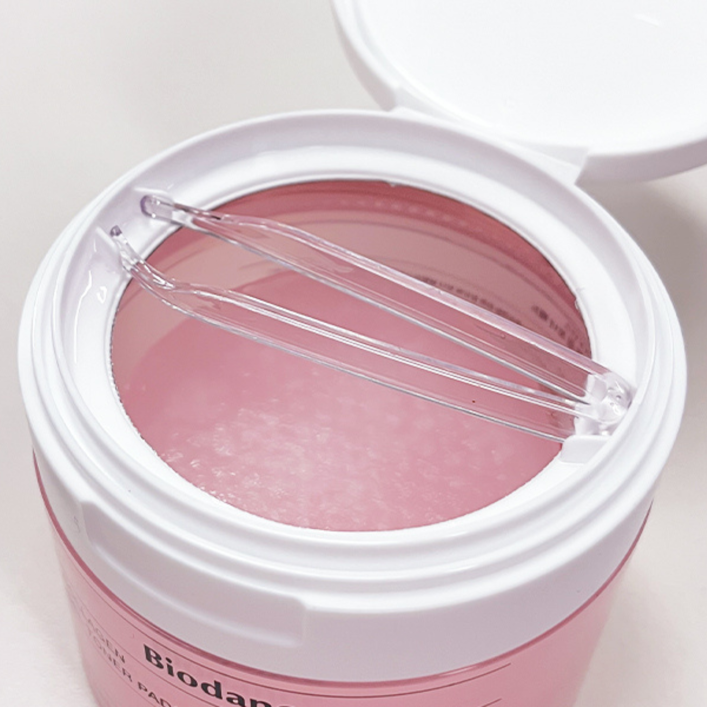 A close-up of pink Biodance Collagen Gel Toner Pad jar with its lid open, showing a translucent pink gel pads inside and a clear plastic spatula resting across the rim. 