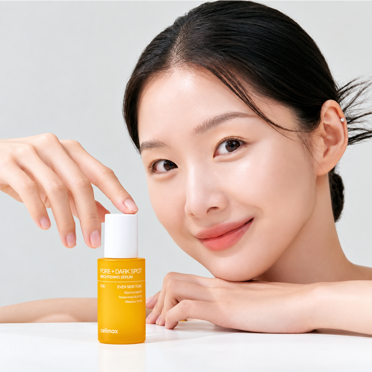 Woman beside a bottle of CELIMAX Pore+Dark Spot Brightening Serum that is resting on a table. She is touching the bottle cap