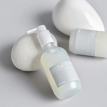 Two bottles of CELIMAX Dual Barrier Mild Gel Cleanser resting on and beside white rocks on a grey background