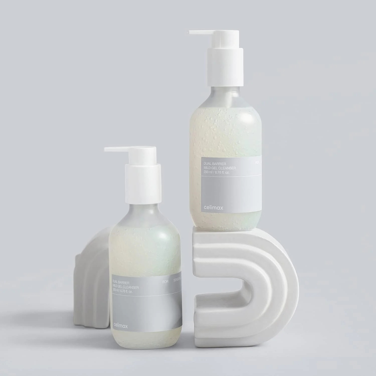Two bottles of CELIMAX Dual Barrier Mild Gel Cleanser on a grey background with rainbow shaped platforms