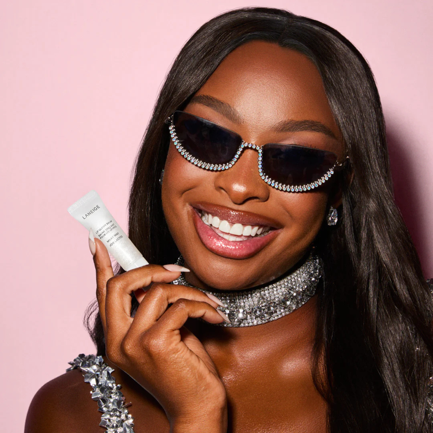Woman wearing sunglasses and a sparkling necklace, holding a LANEIGE Lip Glowy Balm (Milky Way) against a pink background