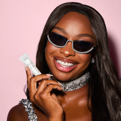 Woman wearing sunglasses and a sparkling necklace, holding a LANEIGE Lip Glowy Balm (Milky Way) against a pink background