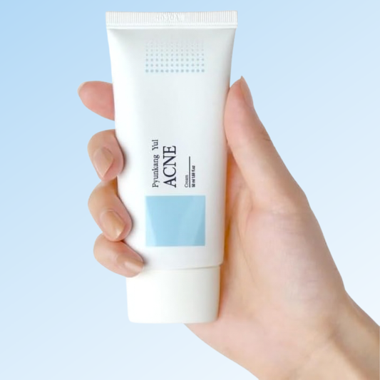 Hand holding a tube of Pyunkang Yul Acne Cream with a blue background