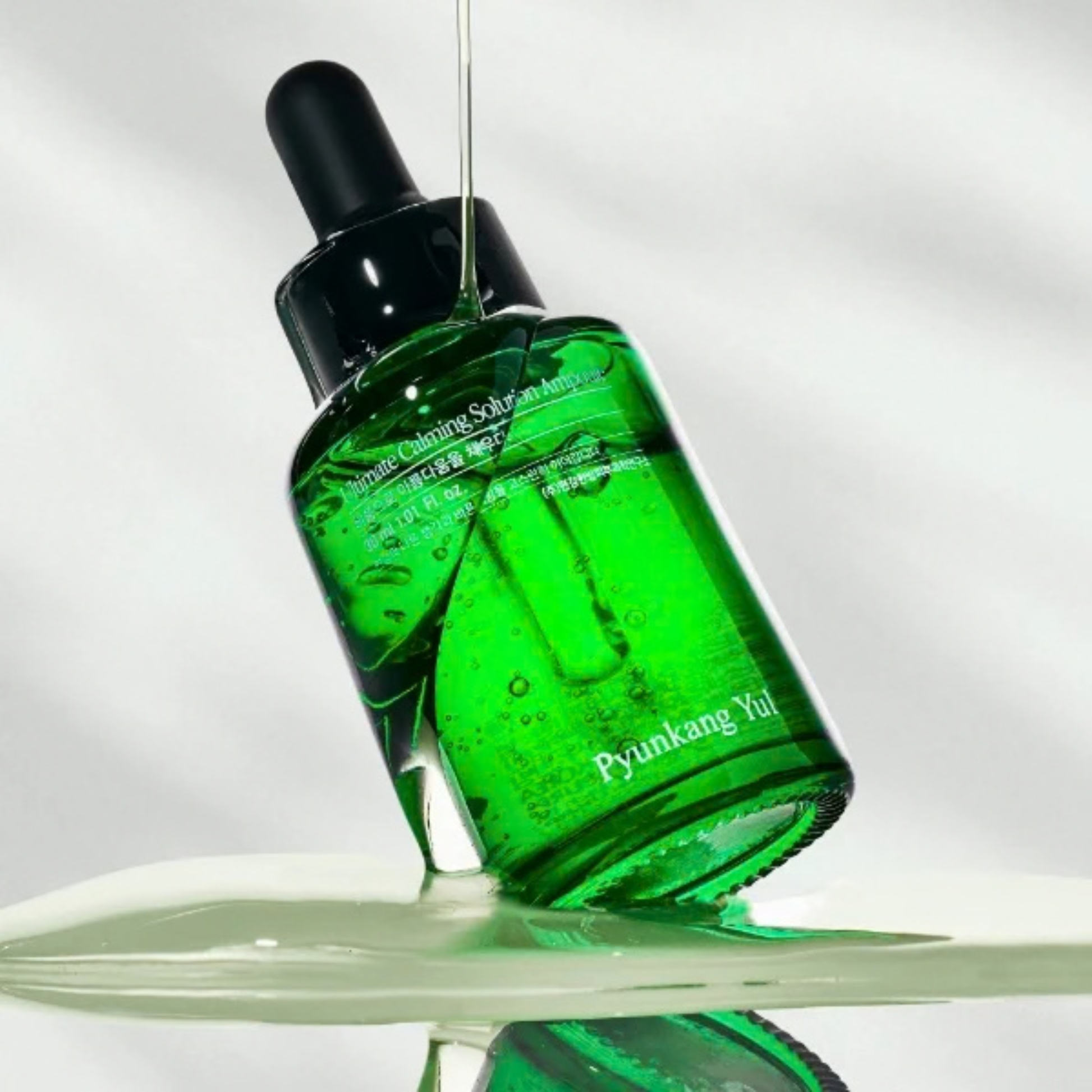 Green when using the Pyunkang Yul Ultimate Calming Solution Ampoule bottle with dropper on a reflective surface with a light gray background. Serum is being poured onto the bottle and pooling on the reflective surface