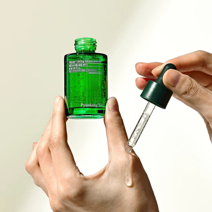Green Pyunkang Yul Ultimate Calming Solution Ampoule bottle with dropper held by a hand while another had is holding the dropper against a neutral background