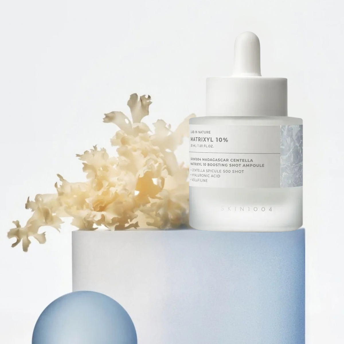 SKIN1004 Lab in Nature Madagascar Centella Matrixyl 10% Boosting Shot Ampoule sits on a pale blue pedestal with dried cream seaweed beside it against a clean white background.