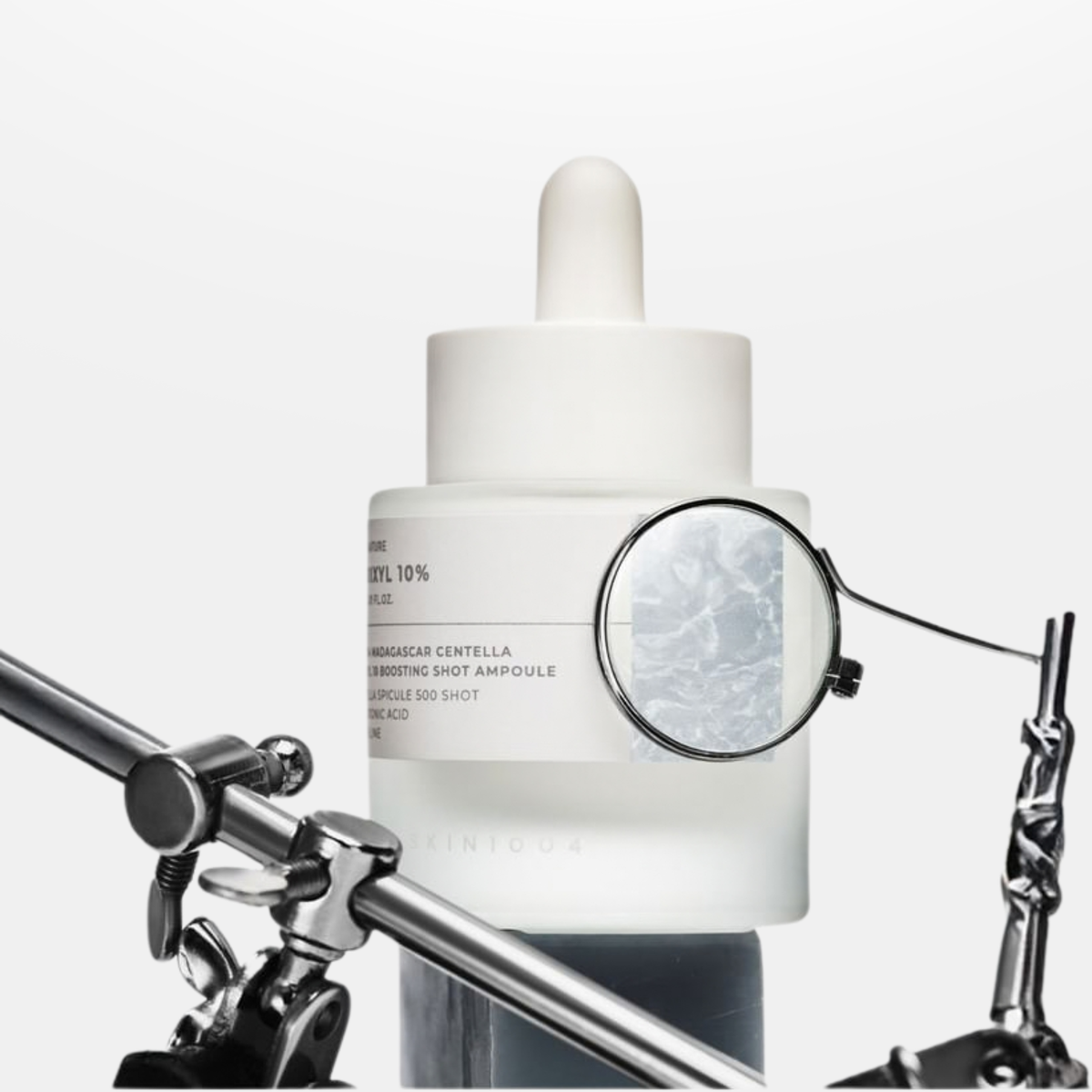 SKIN1004 Lab in Nature Madagascar Centella Matrixyl 10% Boosting Shot Ampoule with a white cap and label, shown against a light gray background with a metal clamp and chain prop framing the lower sides.