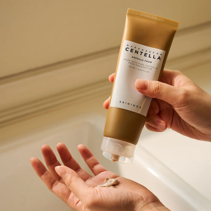 Hand holding and squeezing a tube of SKIN1004 Madagascar Centella Ampoule Foam slightly with some of he product on another hand over a sink.