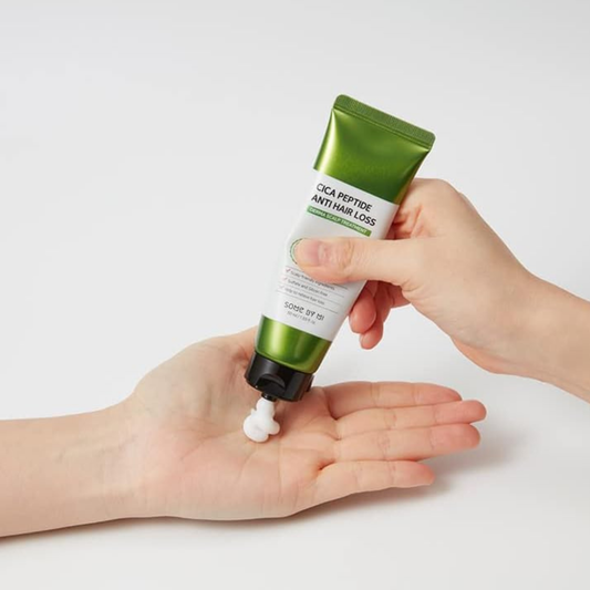 Hand holding a green and white tube labeled SOME BY MI Cica Peptide Anti Hair Loss Derma Scalp Treatment with a small amount of cream being pressed from the tube on a light background.
