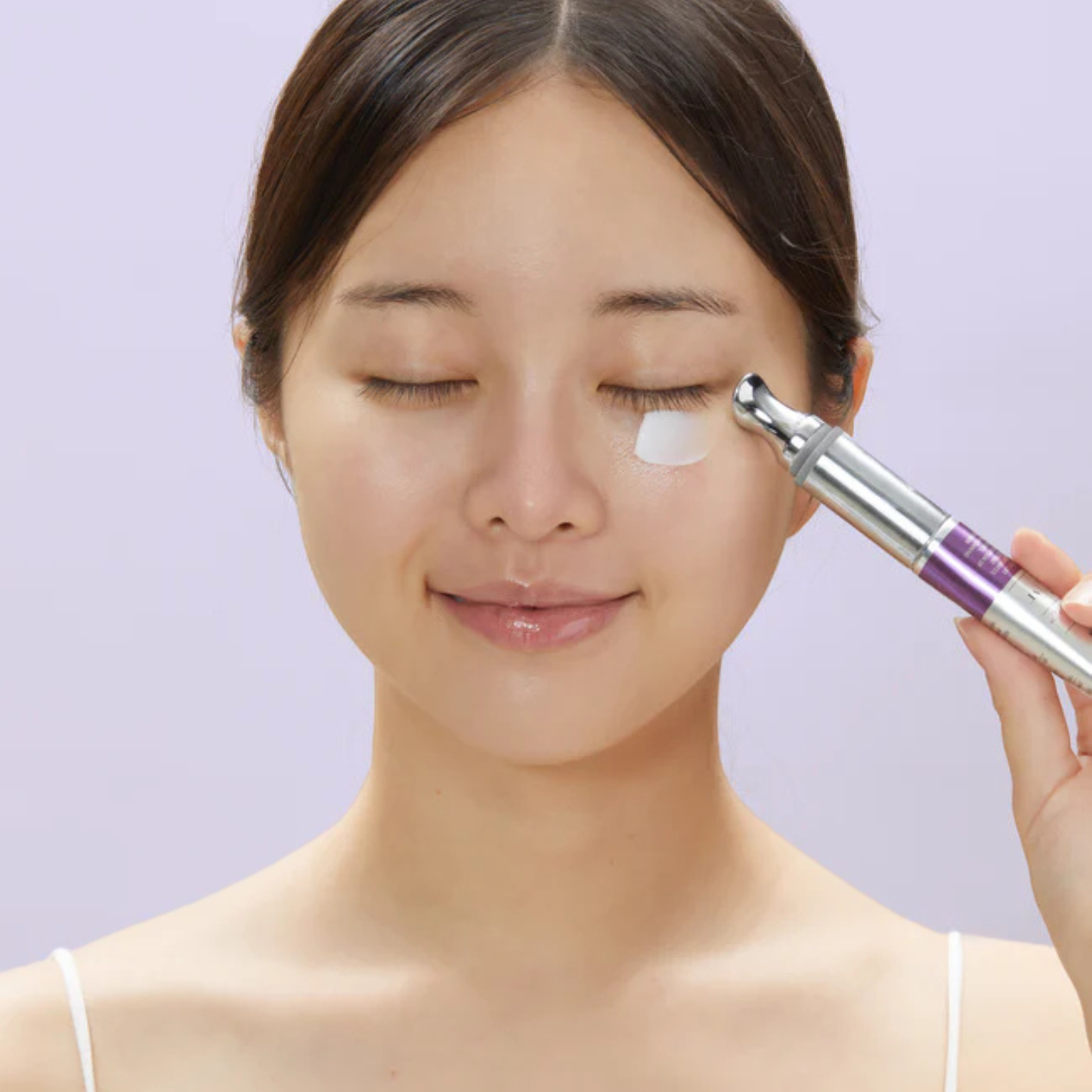 Woman applying VT Cosmetics Reedle Shot Lifting Eye Cream to her face with a the device against a light purple background