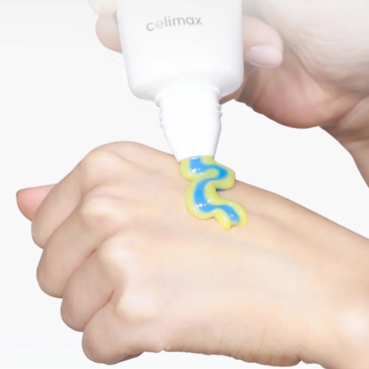 A hand holding a white tube labeled 'celimax' squeezes a squiggle-shaped blue and yellow cream onto the back of another hand, shown against a clean white background.
