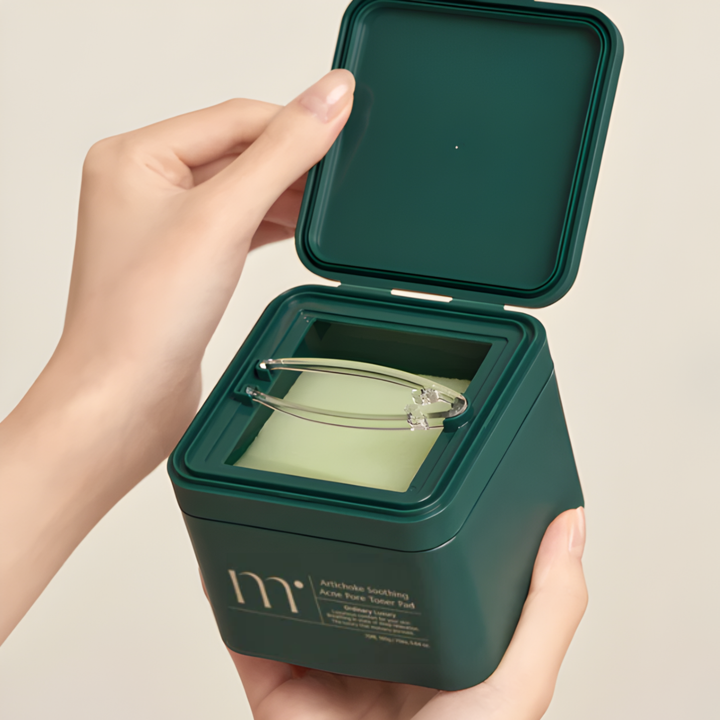 Hand holding a green container of molvany Artichoke Soothing Acne Pore Toner Pads, showing the toner pads and included tool on a beige background