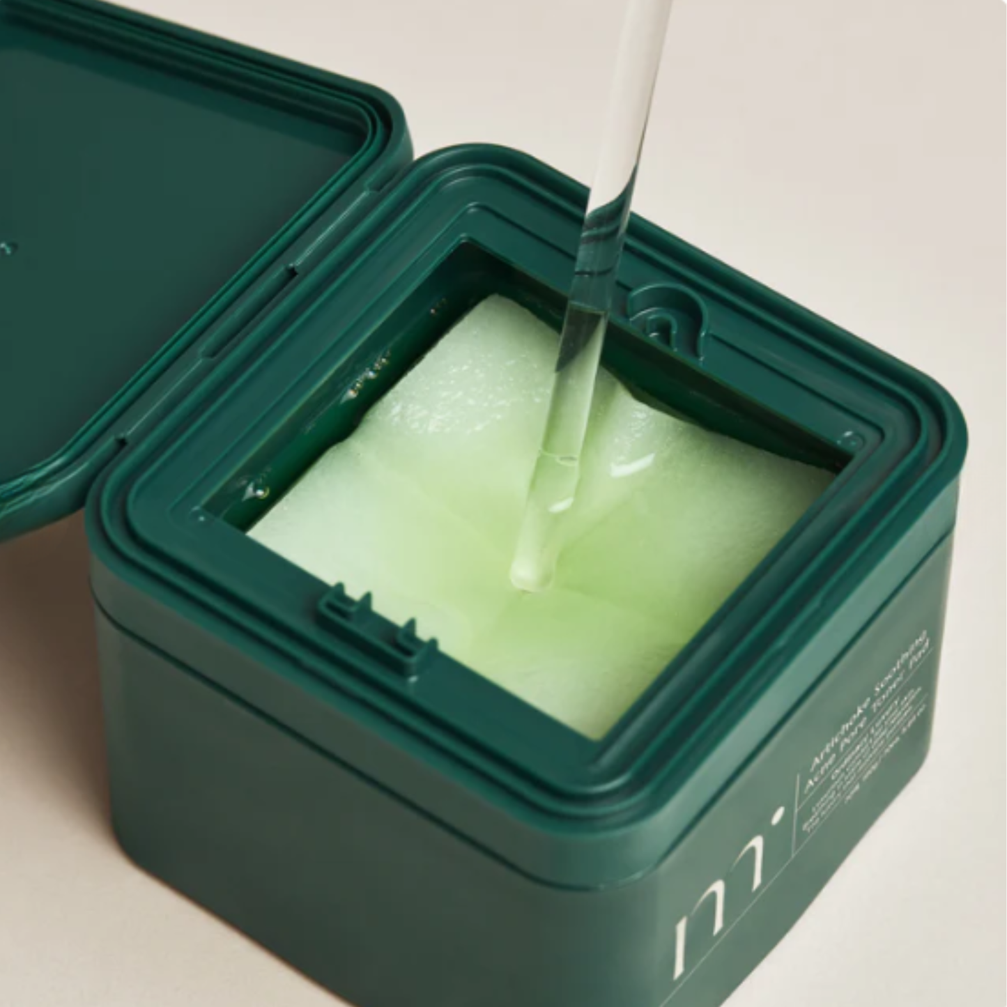 Green container of molvany Artichoke Soothing Acne Pore Toner Pad with liquid being poured  onto the toner pads on a neutral beige background