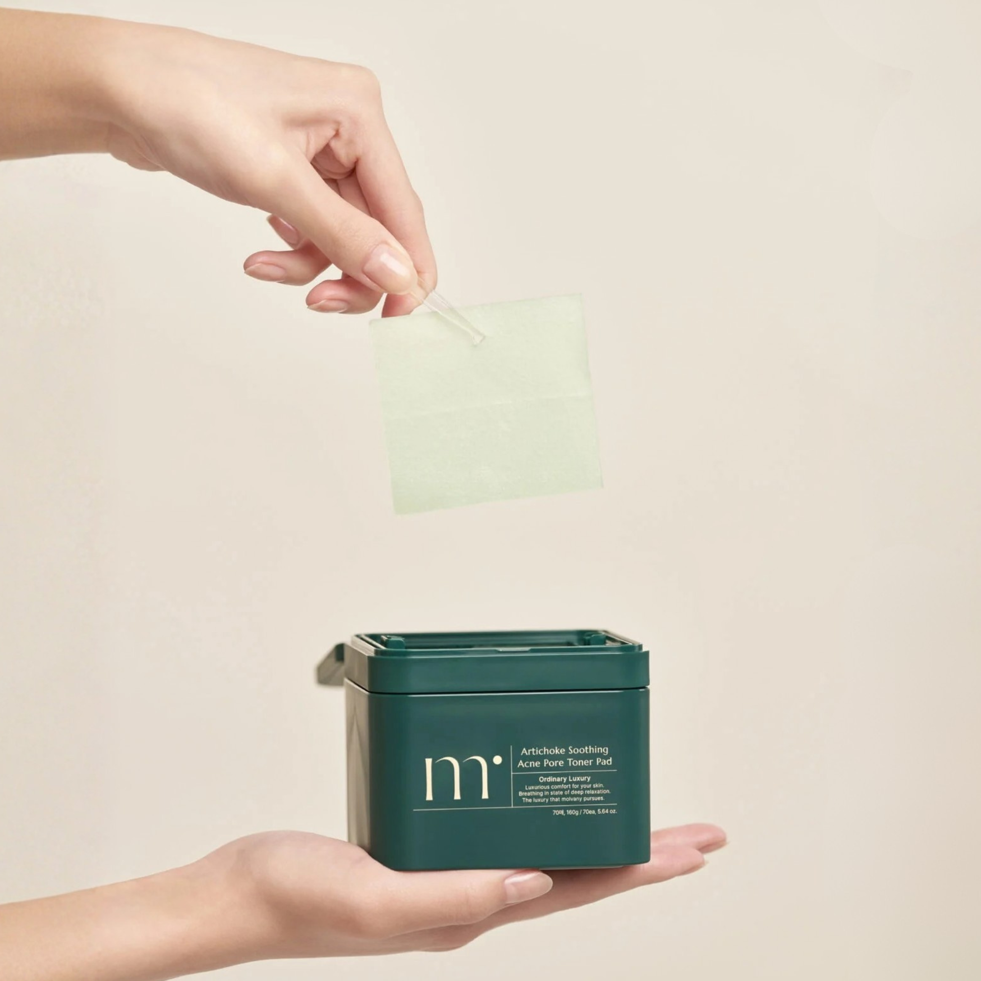 Hand holding a container of molvany Artichoke Soothing Acne Pore Toner Pads with another hand holding a forceps pinching a toner pad on a beige background