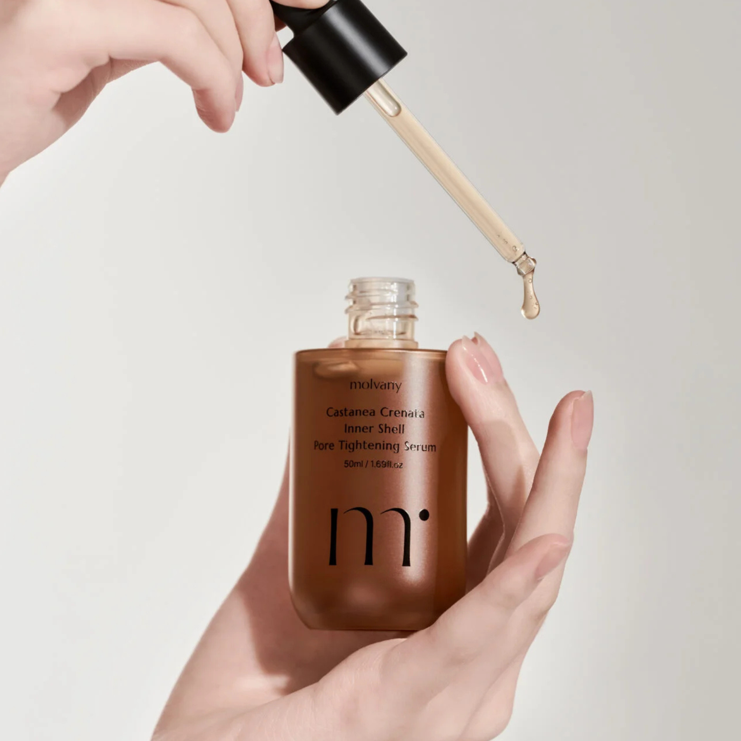 Hand holding a molvany Castanea Crenata Inner Shell Pore Tightening Serum bottle with a dropper, against a plain background